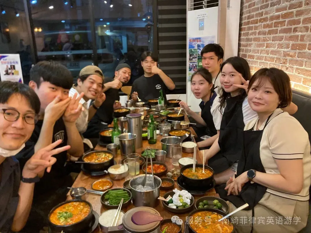 Ten multinational students from Korea, Taiwan, China, and Japan enjoying a dinner together at Miss Jin Sundae restaurant in Angeles’ Koreatown. The students are smiling and posing for a group photo at the table filled with Korean dishes, including Sundae, kimchi, side dishes, soju, and chili peppers. The atmosphere is lively and fun.